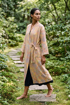 Bata kimono Kihada, jacquard botánico, melocotón: modelo caminando, vista de tres cuartos, en un sendero de piedras en un jardín de musgo