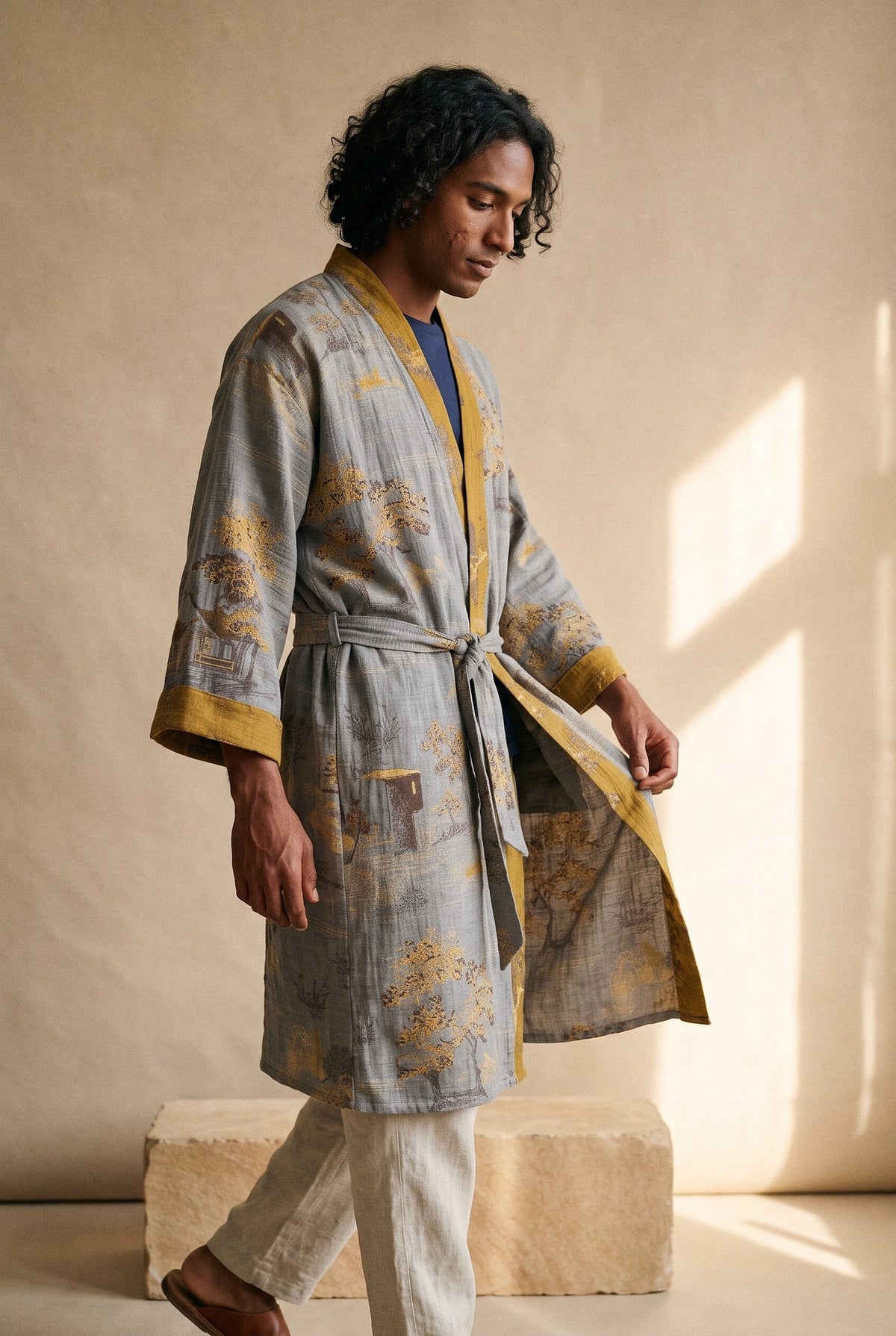 Bata kimono Momiji, jacquard gris, hombre: modelo caminando, en estudio, fondo color piedra y bloque de arenisca
