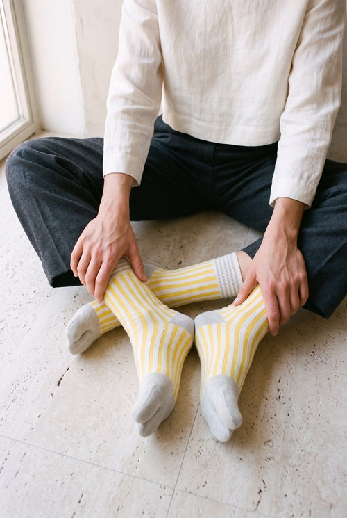 Calcetines tabi Miko, rayas, amarillo: modelo sentada con las piernas cruzadas, plantas a la vista, en una sala desnuda con suelo de travertino