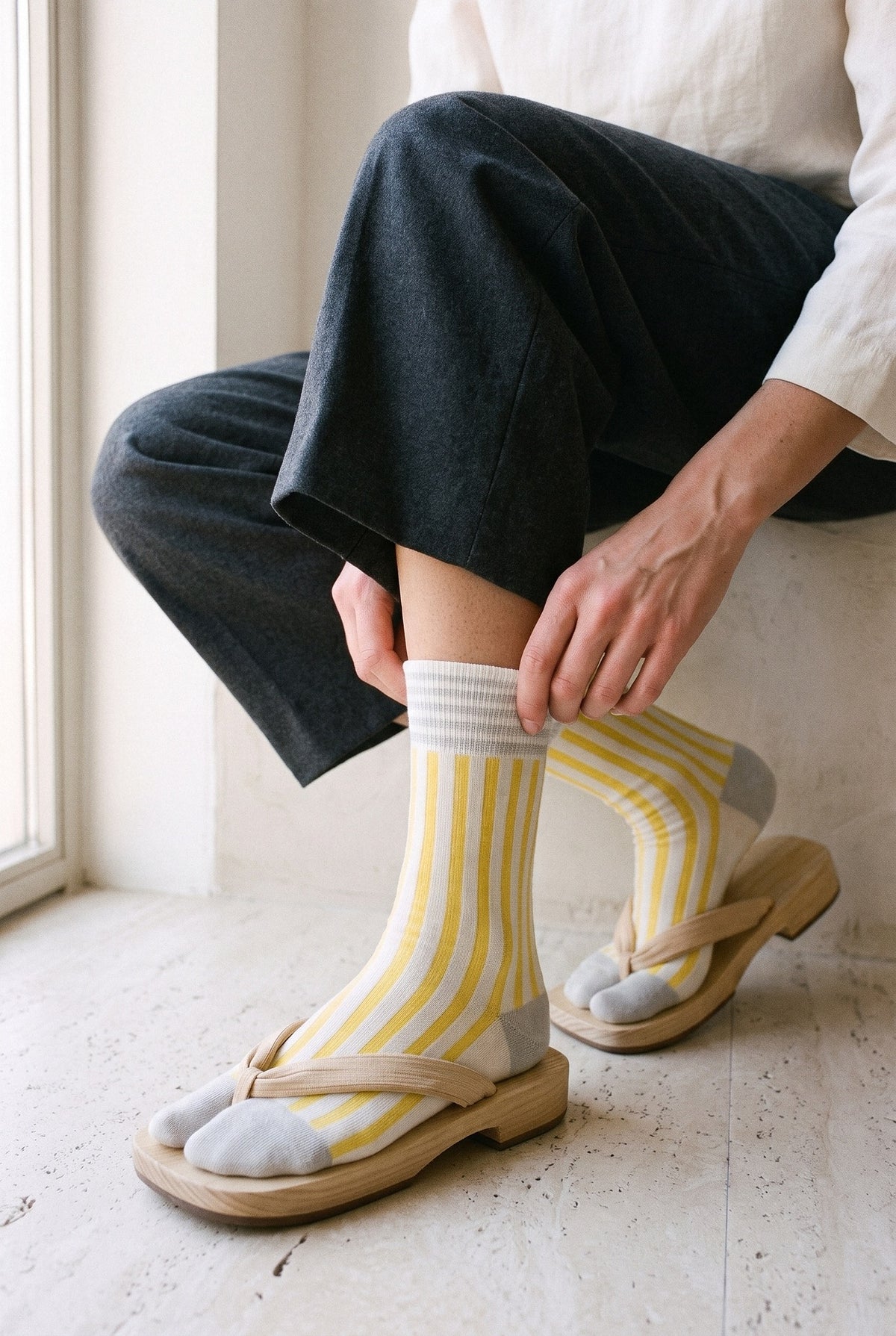 Calcetines tabi Miko, rayas, amarillo: modelo agachada subiéndose un calcetín, en una sala desnuda con suelo de travertino