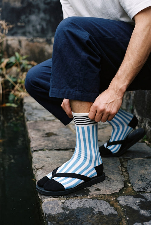Calcetines tabi Miko, rayas, azul: modelo agachado subiéndose un calcetín, junto a un estanque de carpas con piedras oscuras