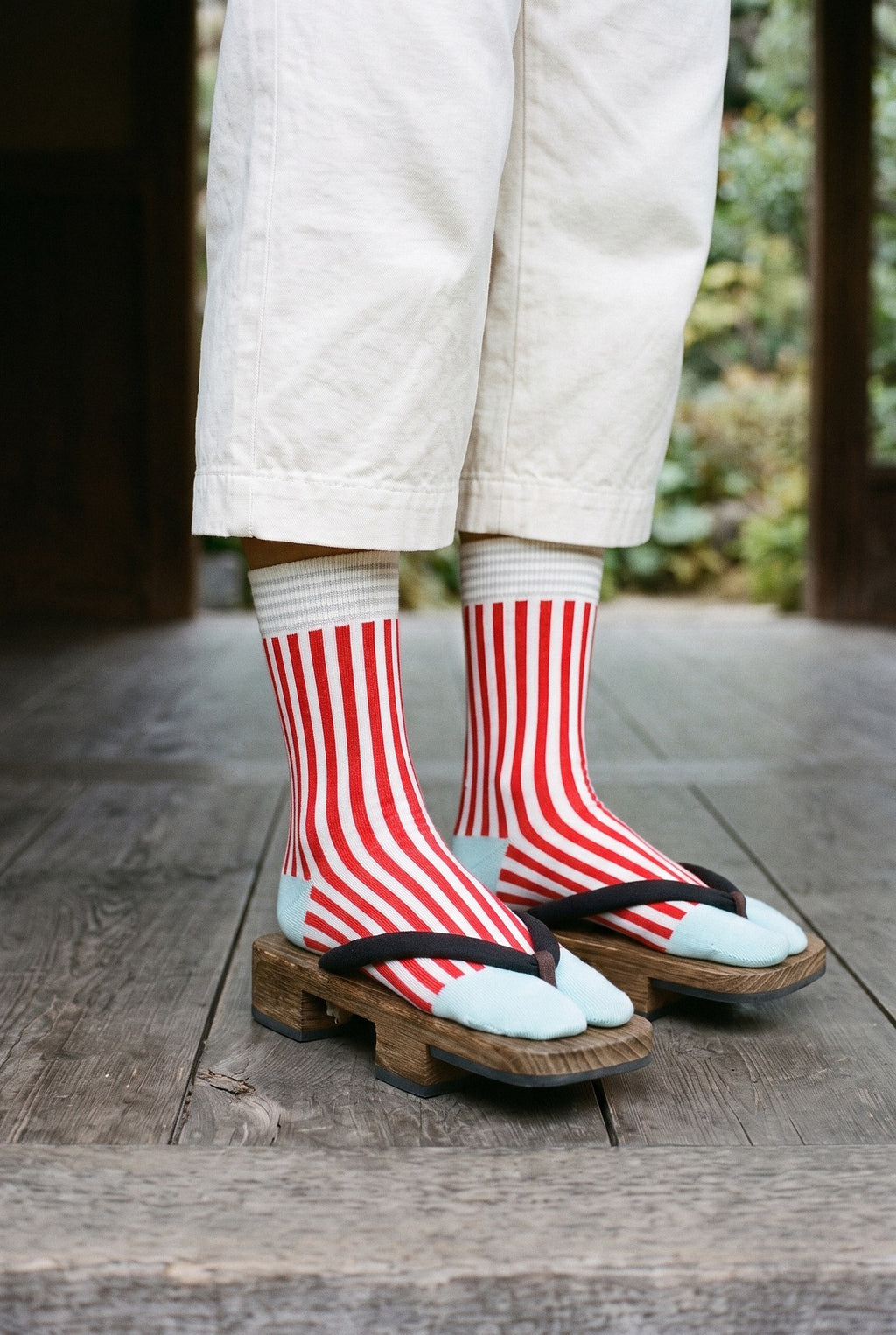 Calcetines tabi Miko, rayas, rojo: detalle de los calcetines, de la rodilla al pie, en un pabellón de madera oscura abierto al jardín