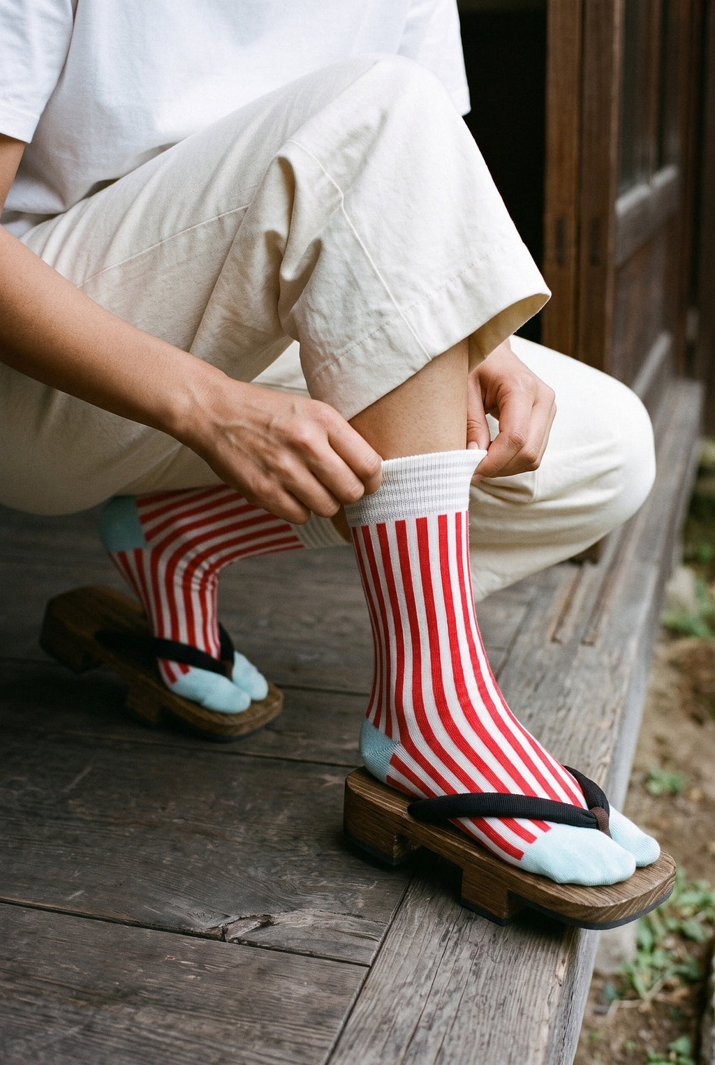 Calcetines tabi Miko, rayas, rojo: modelo agachada subiéndose un calcetín, en un pabellón de madera oscura abierto al jardín