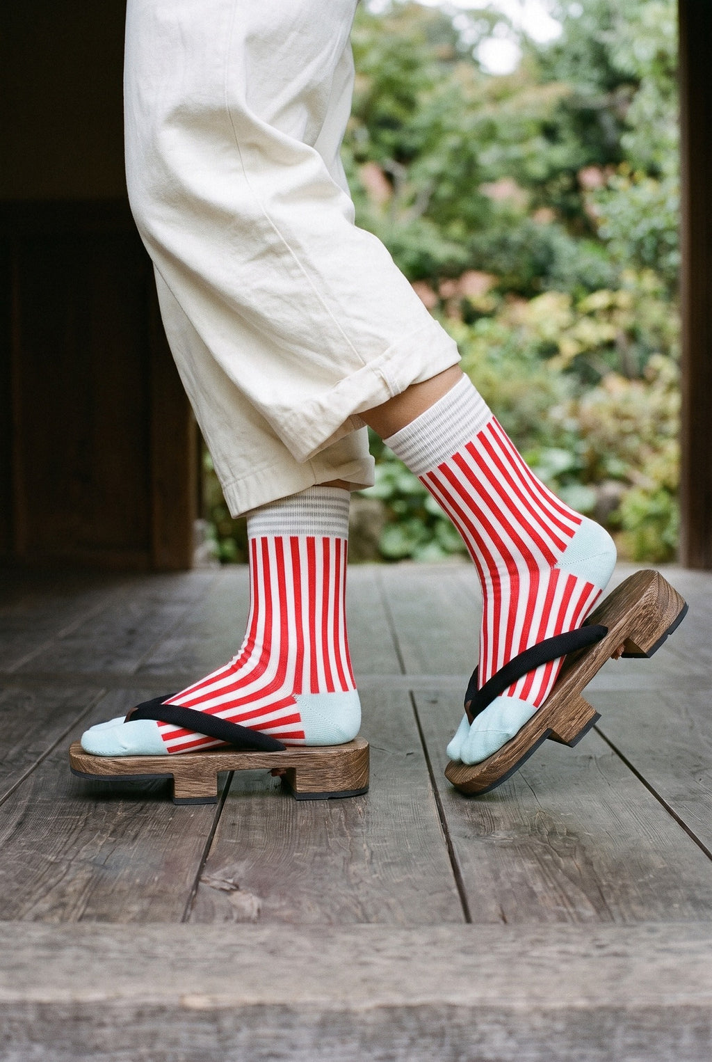 Calcetines tabi Miko, rayas, rojo: detalle de los pies de perfil, en un pabellón de madera oscura abierto al jardín