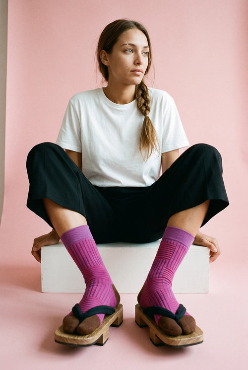 Calcetines tabi Sen, rayas, rosa fucsia: modelo sentada con los tobillos cruzados, en estudio, fondo rosa empolvado con peana blanca