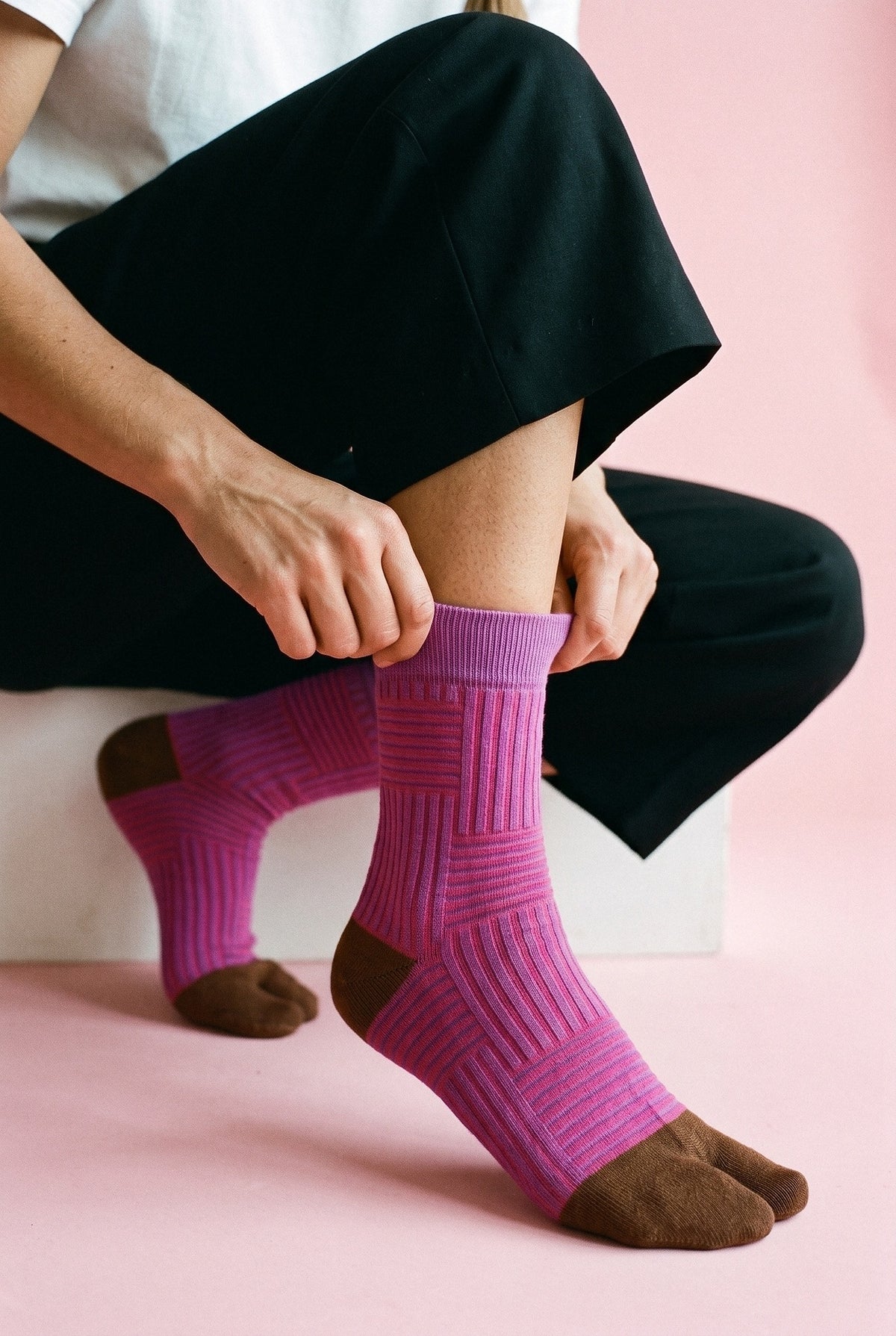 Calcetines tabi Sen, rayas, rosa fucsia: modelo agachada subiéndose un calcetín, en estudio, fondo rosa empolvado con peana blanca