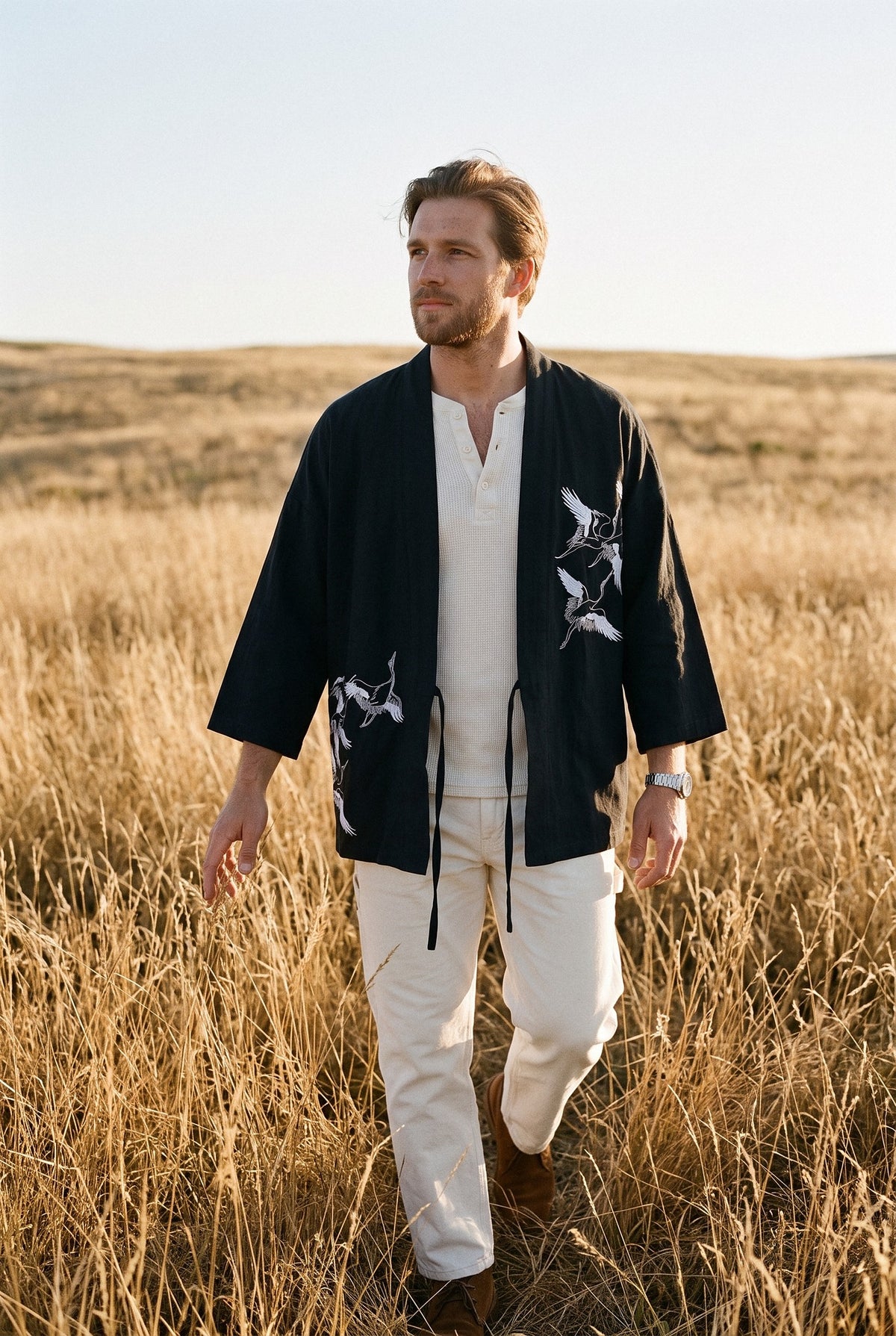 Haori Kaze negro con grullas estampadas, abierto sobre henley y pantalón crudos, en un campo de hierba dorada