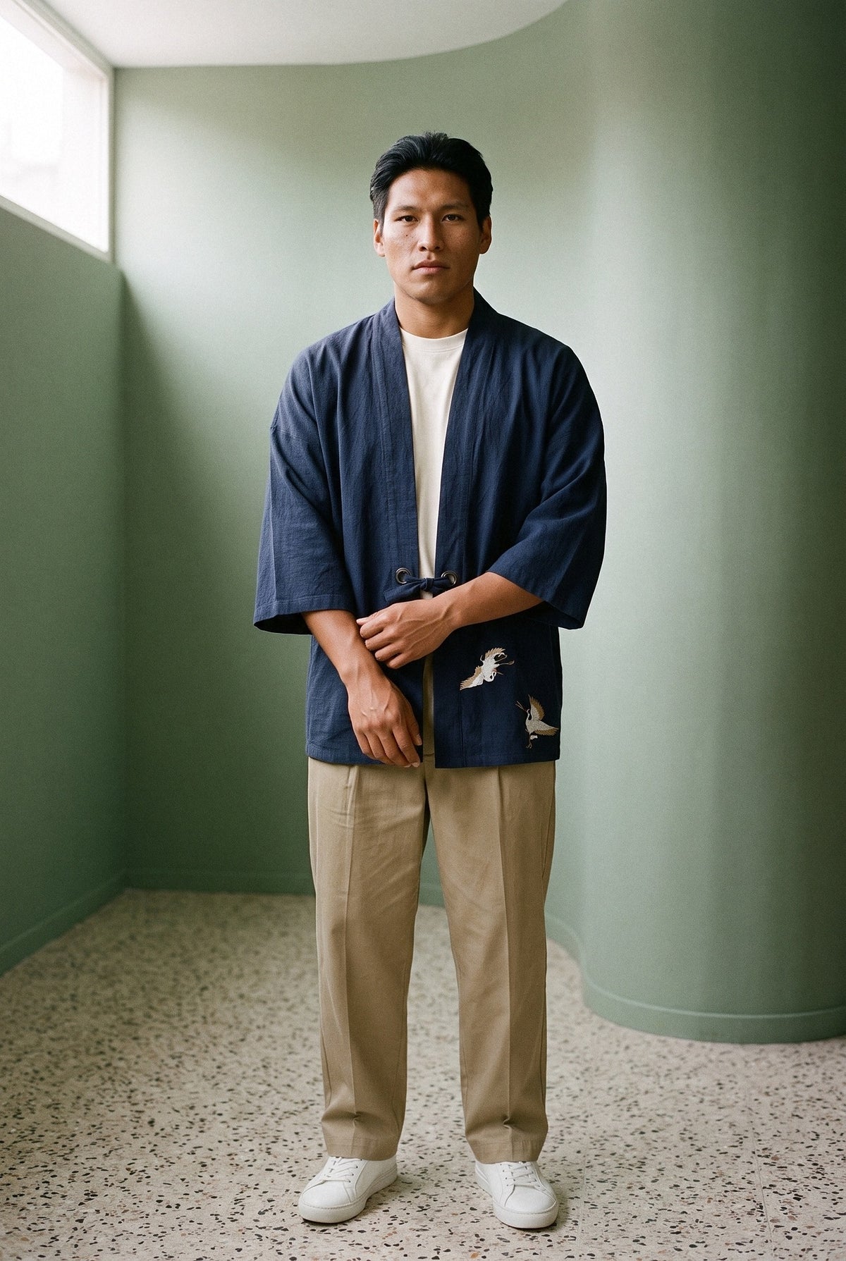 Haori Tsuru azul marino con grullas bordadas, cerrado con su cordón sobre camiseta cruda y pantalón caqui, frente a un muro verde salvia