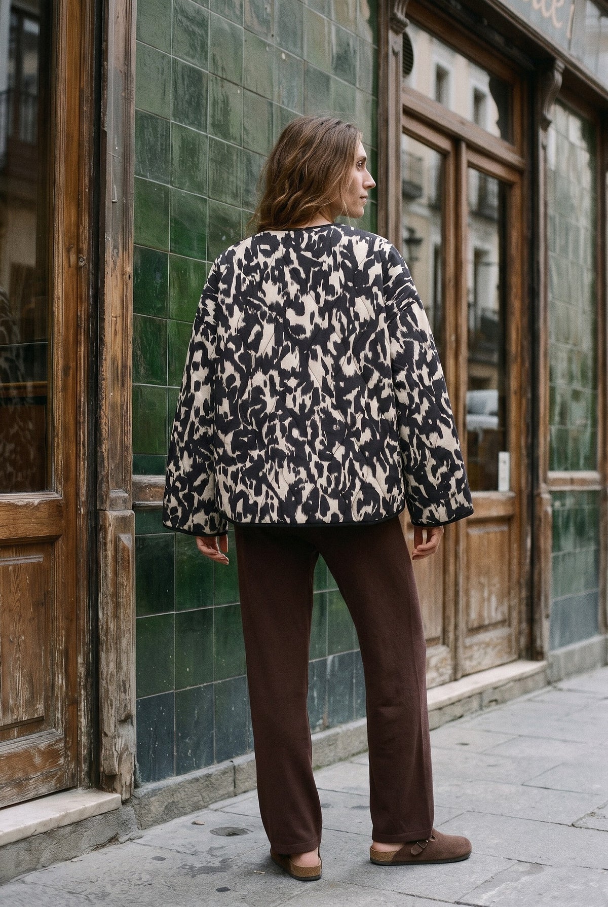Chaqueta acolchada Kokutan, estampado gráfico, negro y blanco: vista de espaldas, ante la fachada de azulejos verdes de un café de Madrid