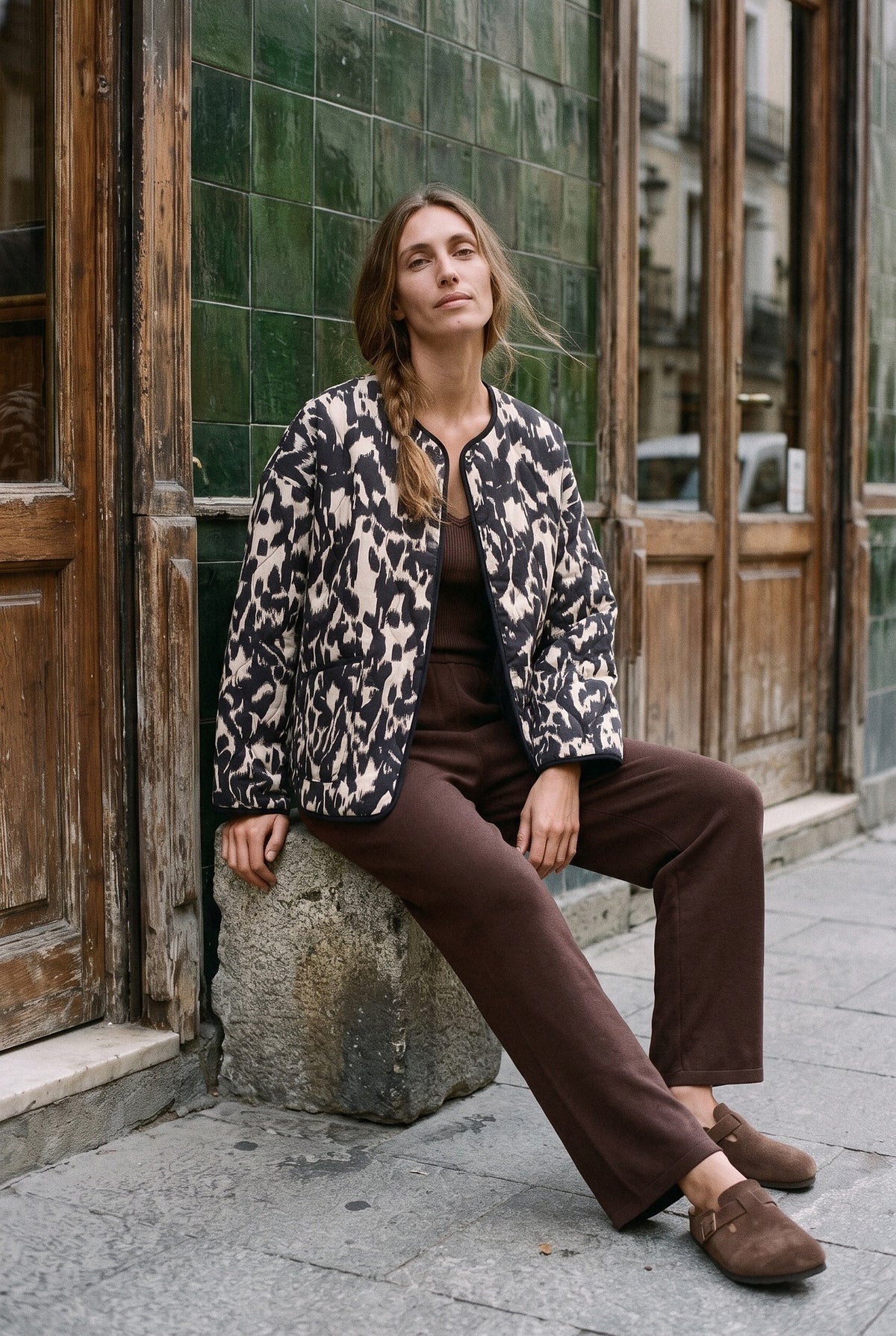 Chaqueta acolchada Kokutan, estampado gráfico, negro y blanco: modelo sentada con una pierna estirada, ante la fachada de azulejos verdes de un café de Madrid