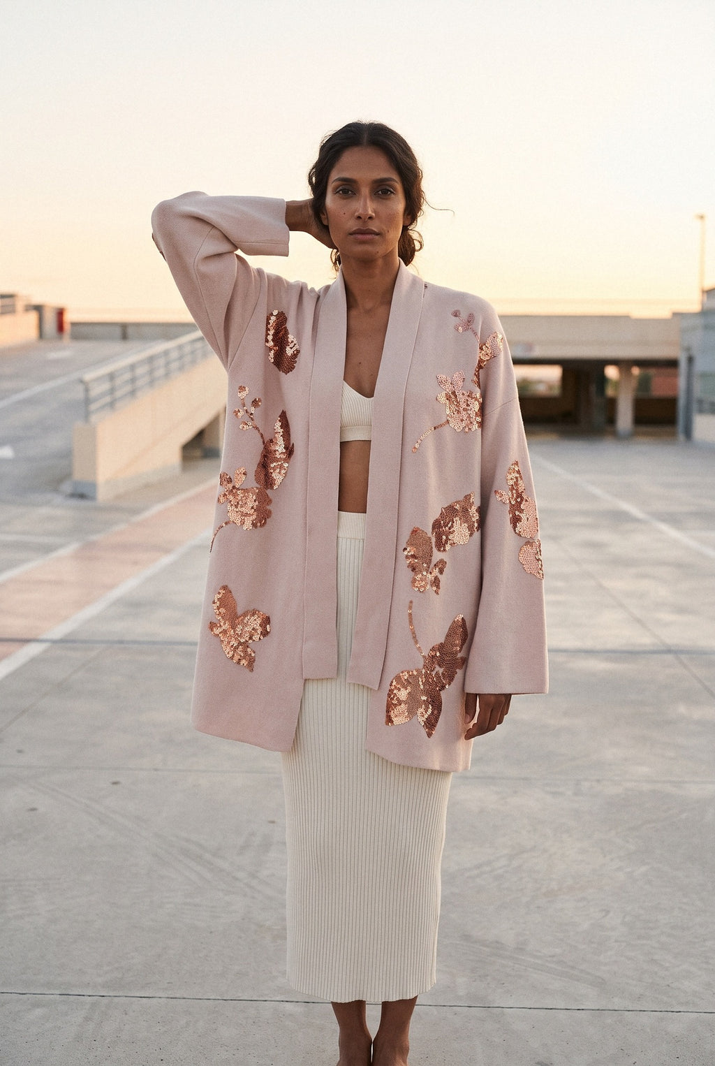 Chaqueta kimono Anzu, flores de lentejuelas, rosa empolvado: modelo con una mano en la nuca, en la azotea de un aparcamiento de hormigón claro