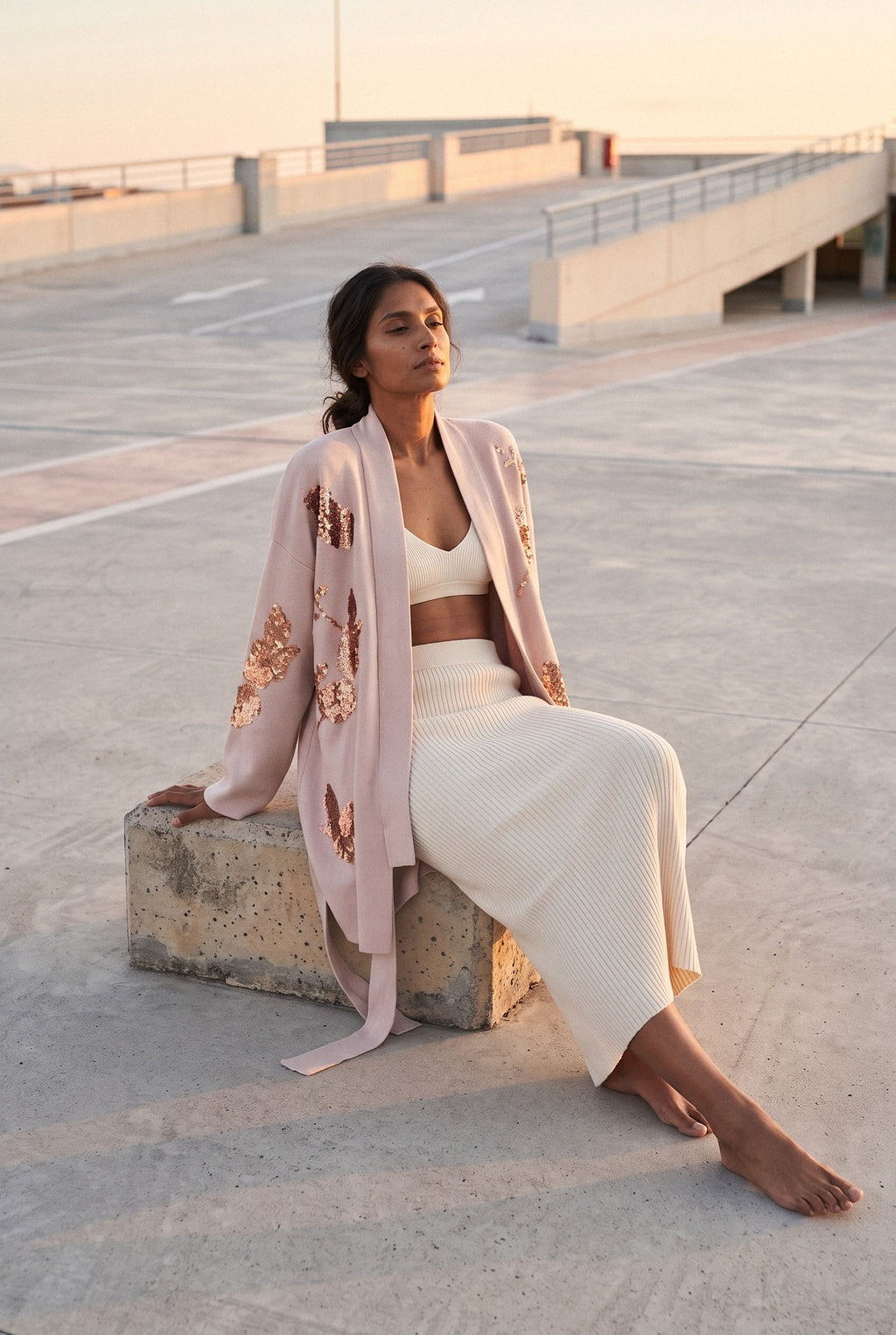 Chaqueta kimono Anzu, flores de lentejuelas, rosa empolvado: modelo sentada con una pierna estirada, en la azotea de un aparcamiento de hormigón claro
