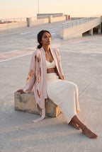 Chaqueta kimono Anzu, flores de lentejuelas, rosa empolvado: modelo sentada con una pierna estirada, en la azotea de un aparcamiento de hormigón claro