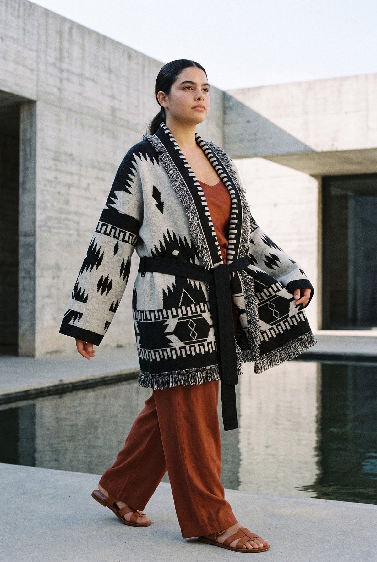 Chaqueta kimono Azteca, cachemira jacquard con flecos: modelo caminando, en un patio de hormigón visto con lámina de agua