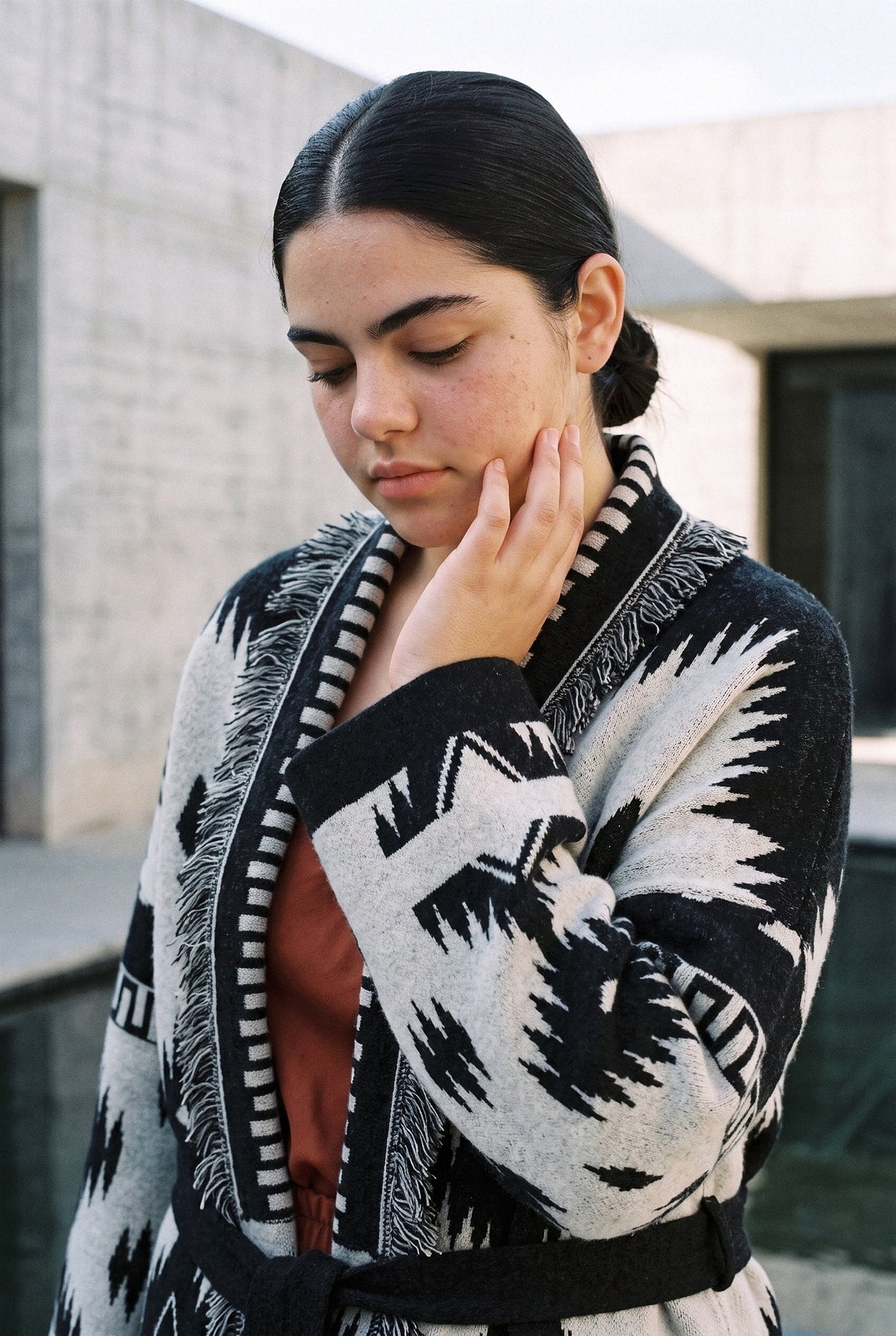 Chaqueta kimono Azteca, cachemira jacquard con flecos: retrato, mano en la barbilla, en un patio de hormigón visto con lámina de agua
