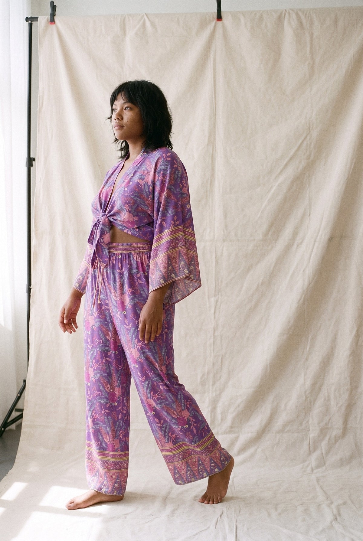 Conjunto kimono Murasaki, kimono corto y pantalón, morado: modelo caminando, vista de tres cuartos, en estudio, fondo de lienzo crudo