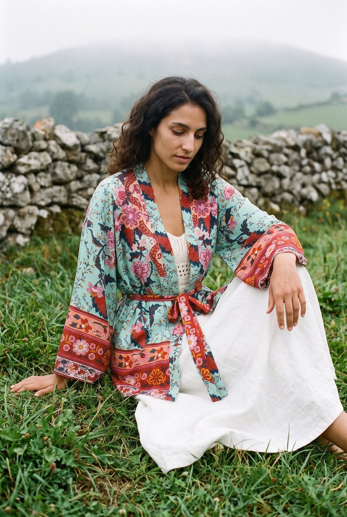 Kimono corto Aoba, pavo real y flores, verde: modelo sentada en el suelo, en un prado asturiano con niebla y muro de piedra seca