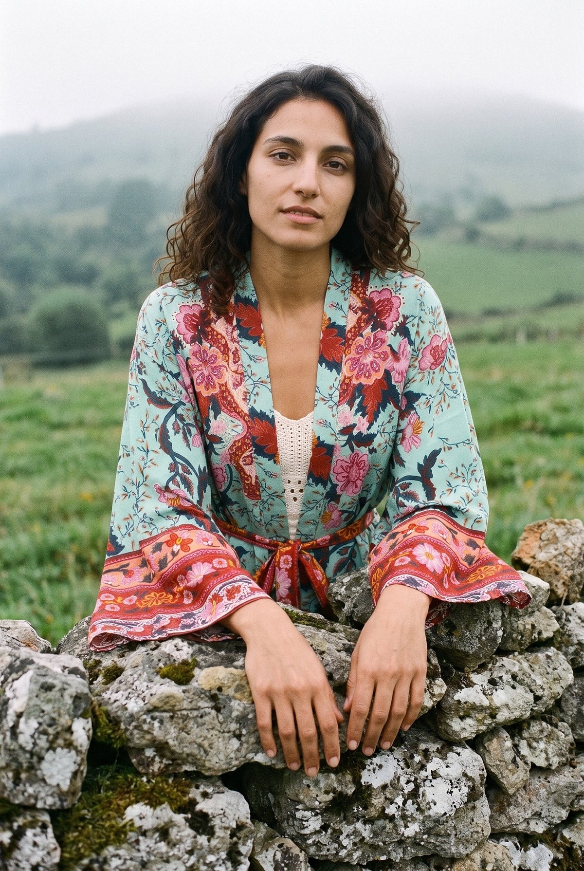 Kimono corto Aoba, pavo real y flores, verde: modelo apoyada en un muro, en un prado asturiano con niebla y muro de piedra seca