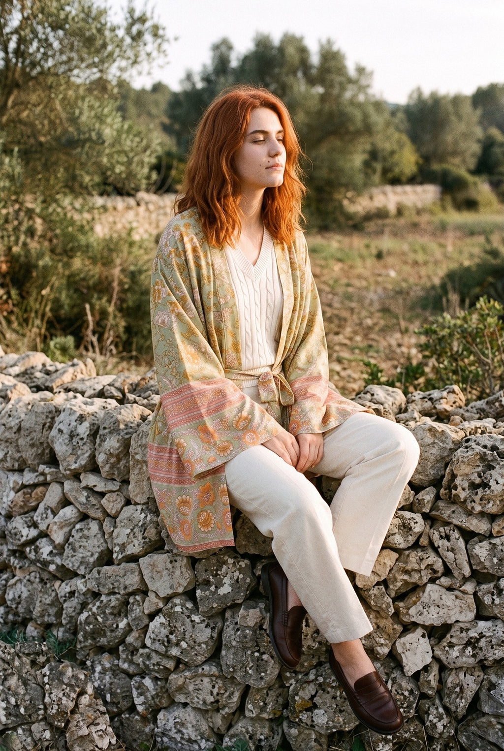 Kimono corto Asahi, flores, verde salvia: modelo sentada de lado con las piernas cruzadas, junto a un muro de piedra seca en Mallorca
