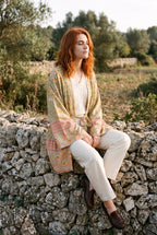 Kimono corto Asahi, flores, verde salvia: modelo sentada de lado con las piernas cruzadas, junto a un muro de piedra seca en Mallorca