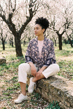 Kimono corto Hai, flores, azul marino y rosa: modelo sentada de lado con las piernas cruzadas, en un almendral en flor de Mallorca