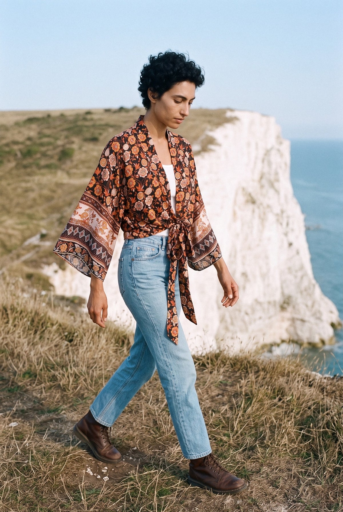 Kimono corto Kaki, flores, negro y teja: modelo caminando, vista de tres cuartos, en lo alto de acantilados blancos frente al mar