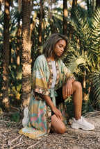 Kimono largo Koke, pavo real y flores, verde salvia: modelo con una rodilla en el suelo, en un palmeral de Elche