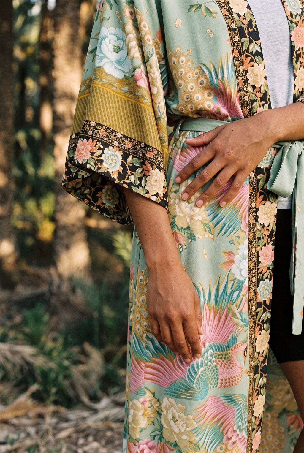 Kimono largo Koke, pavo real y flores, verde salvia: detalle de la manga y del estampado, en un palmeral de Elche