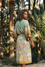 Kimono largo Koke, pavo real y flores, verde salvia: modelo de espaldas, mirando por encima del hombro, en un palmeral de Elche