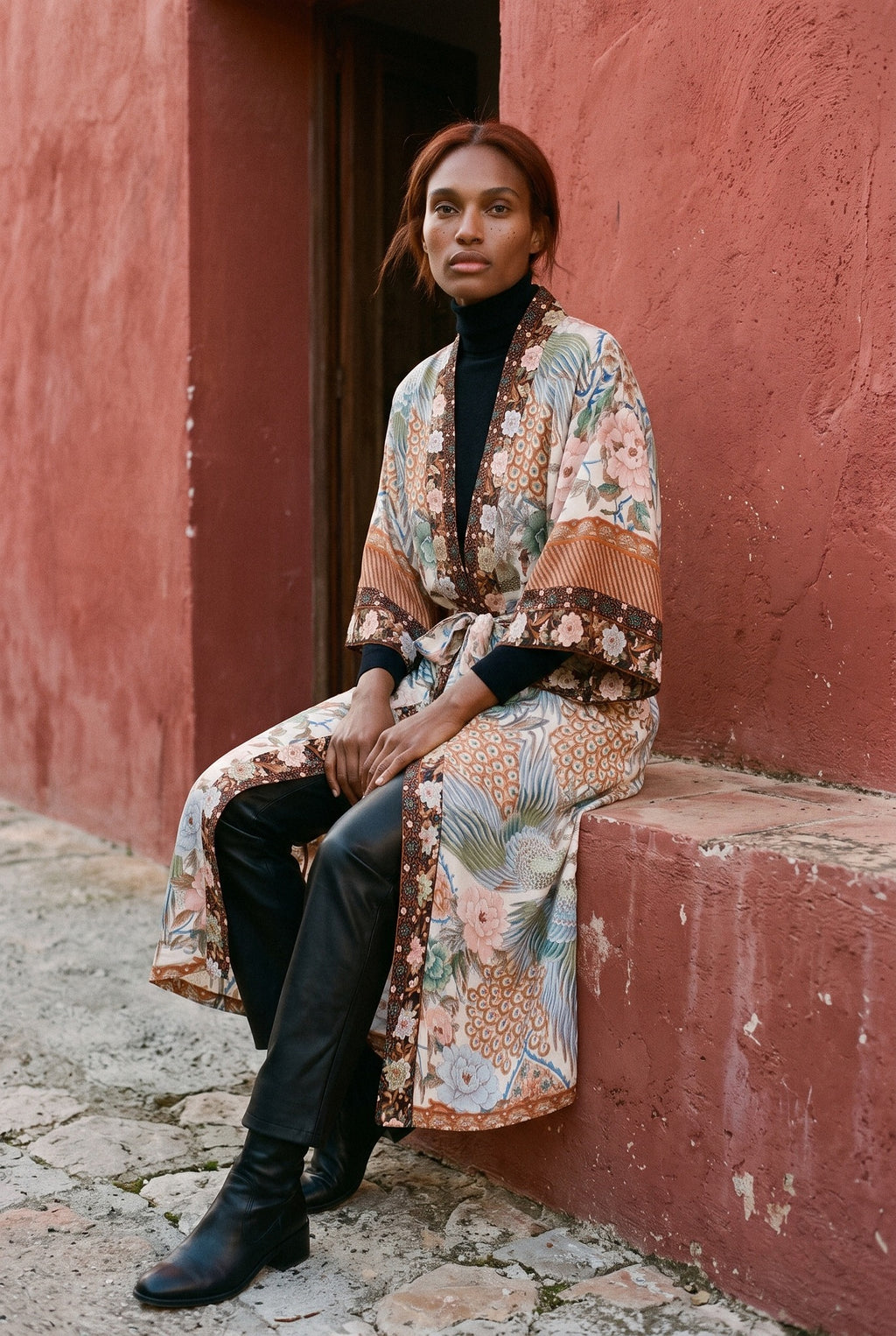 Kimono largo Kuri, pavo real y flores, crudo y ámbar: modelo sentada de lado con las piernas cruzadas, ante un muro de estuco rojo ocre de un pueblo mediterráneo