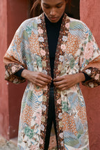 Kimono largo Kuri, pavo real y flores, crudo y ámbar: detalle del estampado y del tejido, ante un muro de estuco rojo ocre de un pueblo mediterráneo
