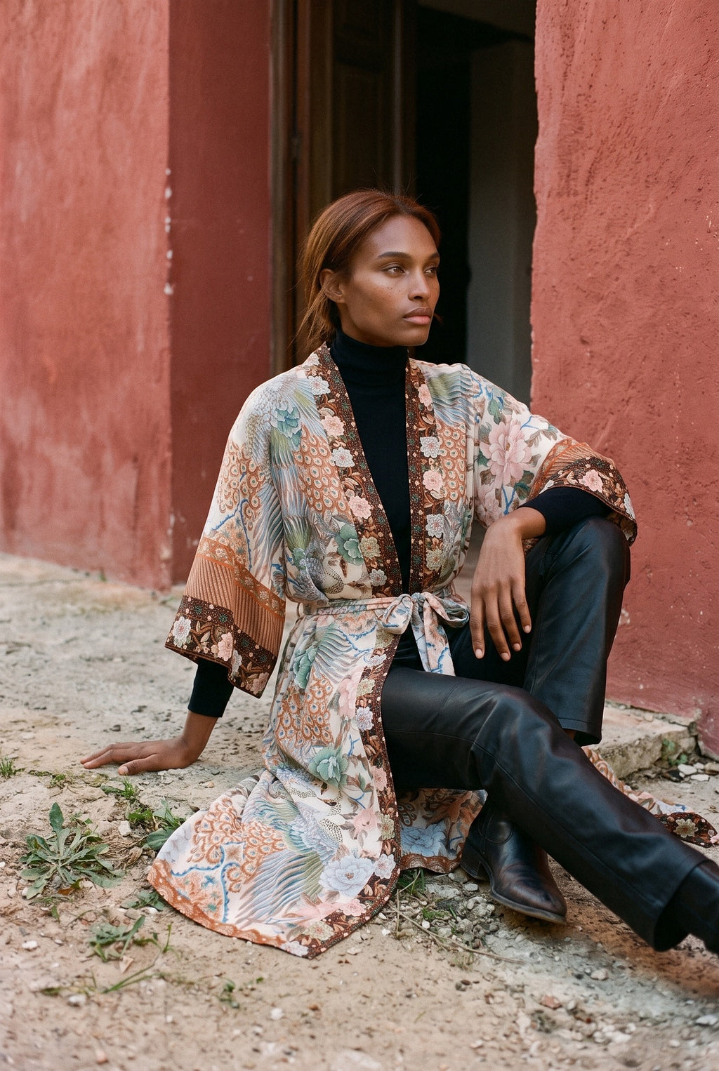 Kimono largo Kuri, pavo real y flores, crudo y ámbar: modelo sentada en el suelo, ante un muro de estuco rojo ocre de un pueblo mediterráneo