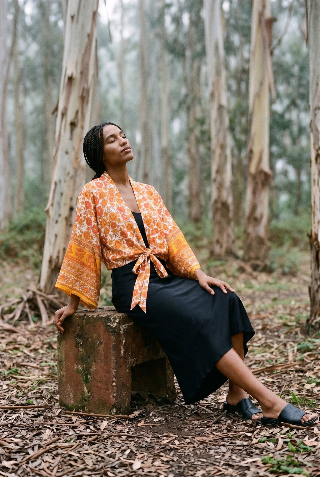 Kimono corto Yamabuki, flores, marfil y naranja: modelo sentada con una pierna estirada, en un bosque de eucaliptos de Cantabria