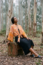 Kimono corto Yamabuki, flores, marfil y naranja: modelo sentada con una pierna estirada, en un bosque de eucaliptos de Cantabria