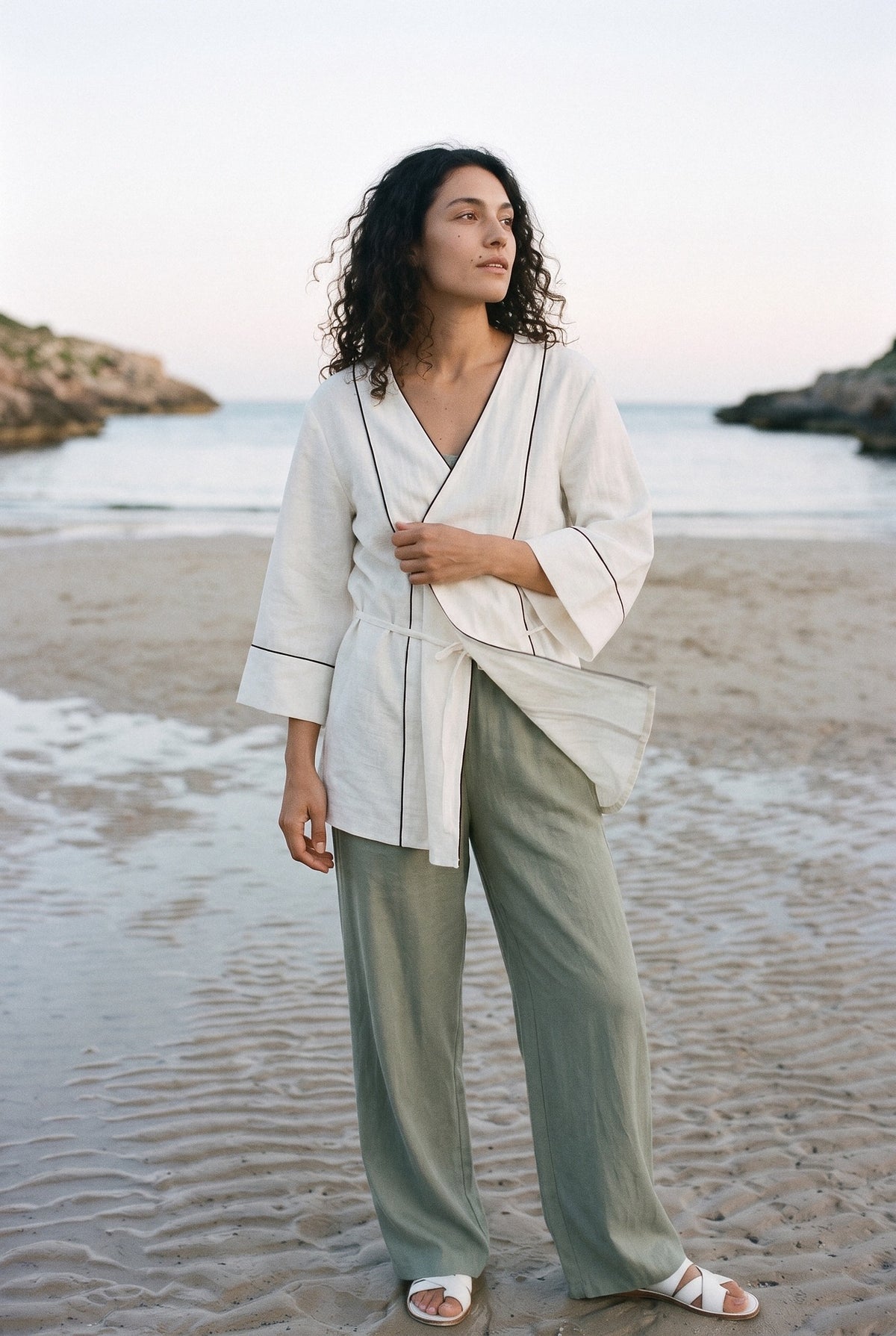 Kimono corto Yuki, blusa cruzada, blanco: modelo con el tejido movido por la brisa, en una cala de arena de Menorca