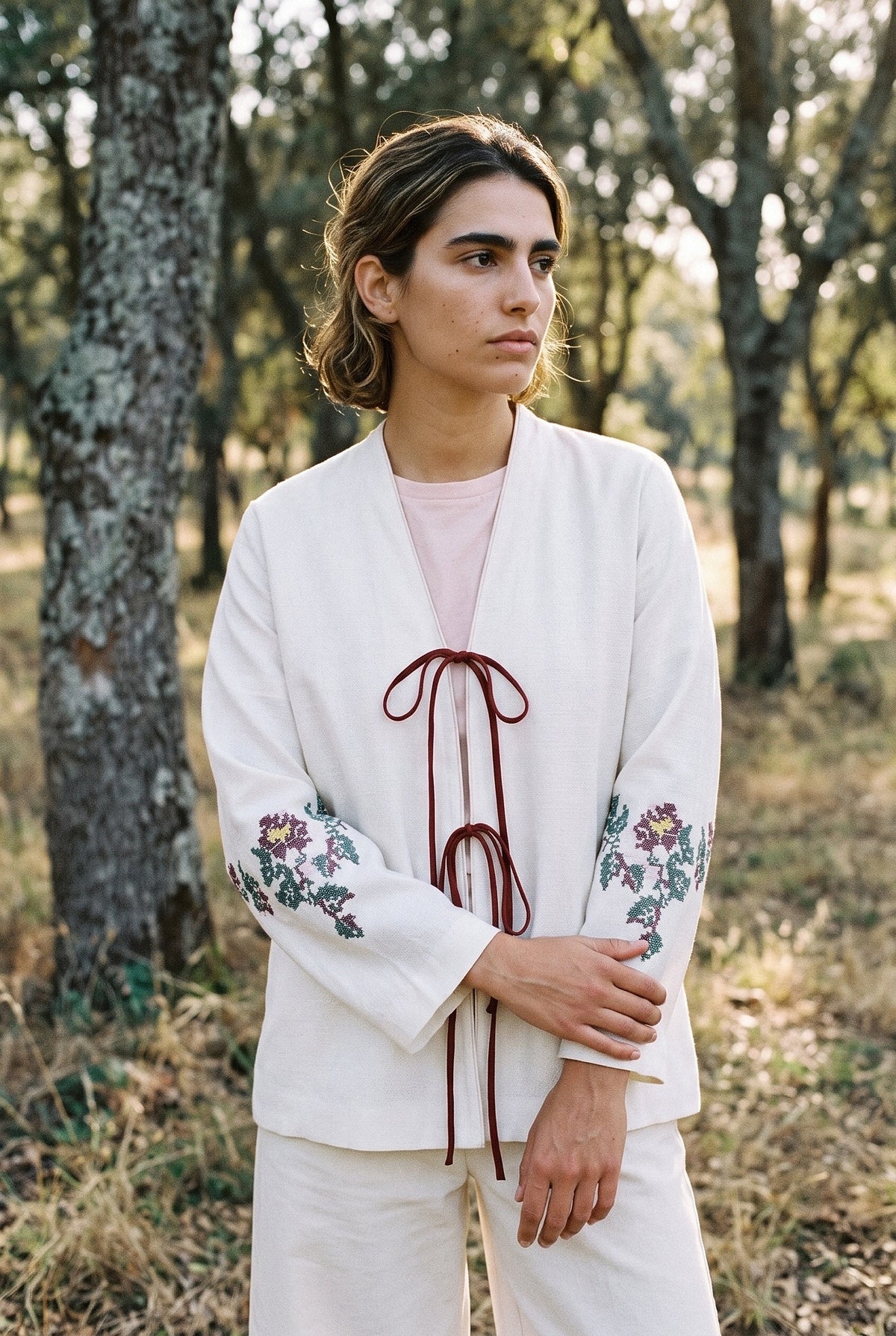 Kimono corto Yuri, bordado floral, crudo: modelo con los brazos cruzados, en un alcornocal de Extremadura