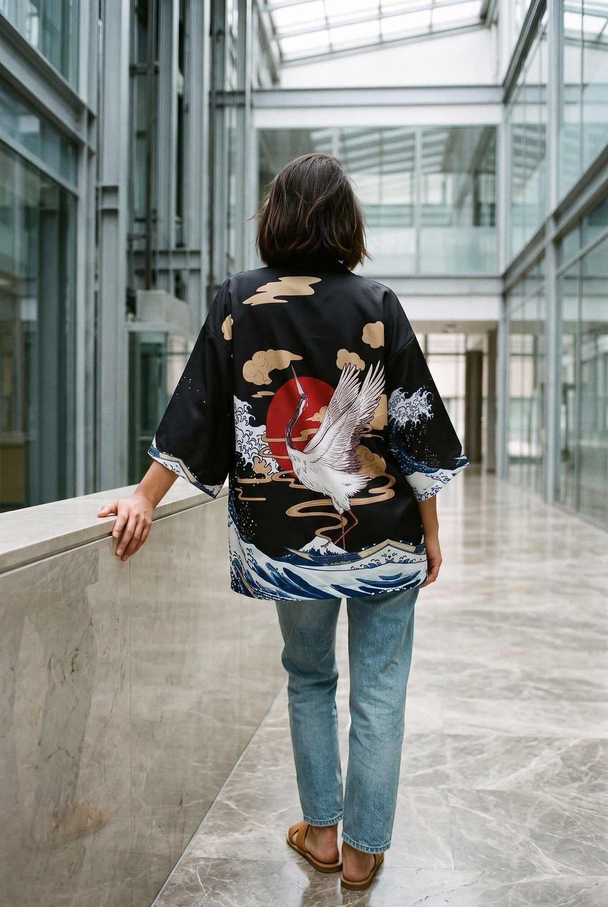 Kimono japonés Tanchō, grullas danzantes, negro: modelo de espaldas, apoyada en un muro, en un atrio de cristal y acero con suelo de mármol