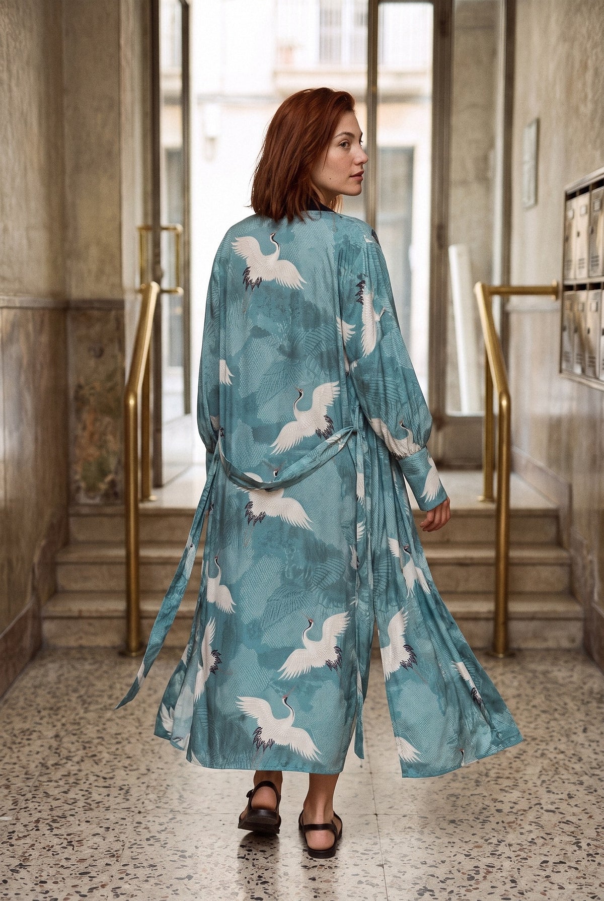 Kimono largo Aoi, estampado turquesa, mujer: modelo de espaldas, alejándose despacio, en un vestíbulo de terrazo de los años 60 en Barcelona