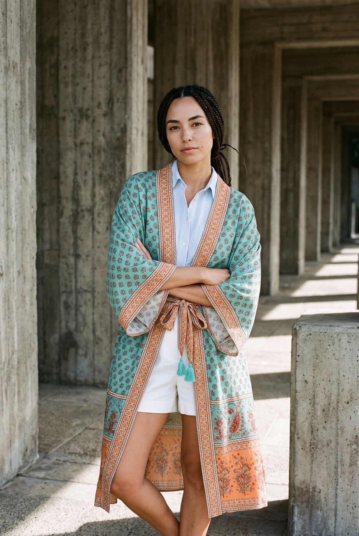Kimono largo Nagisa, cenefas y flores, turquesa: modelo con los brazos cruzados, bajo una columnata brutalista de hormigón