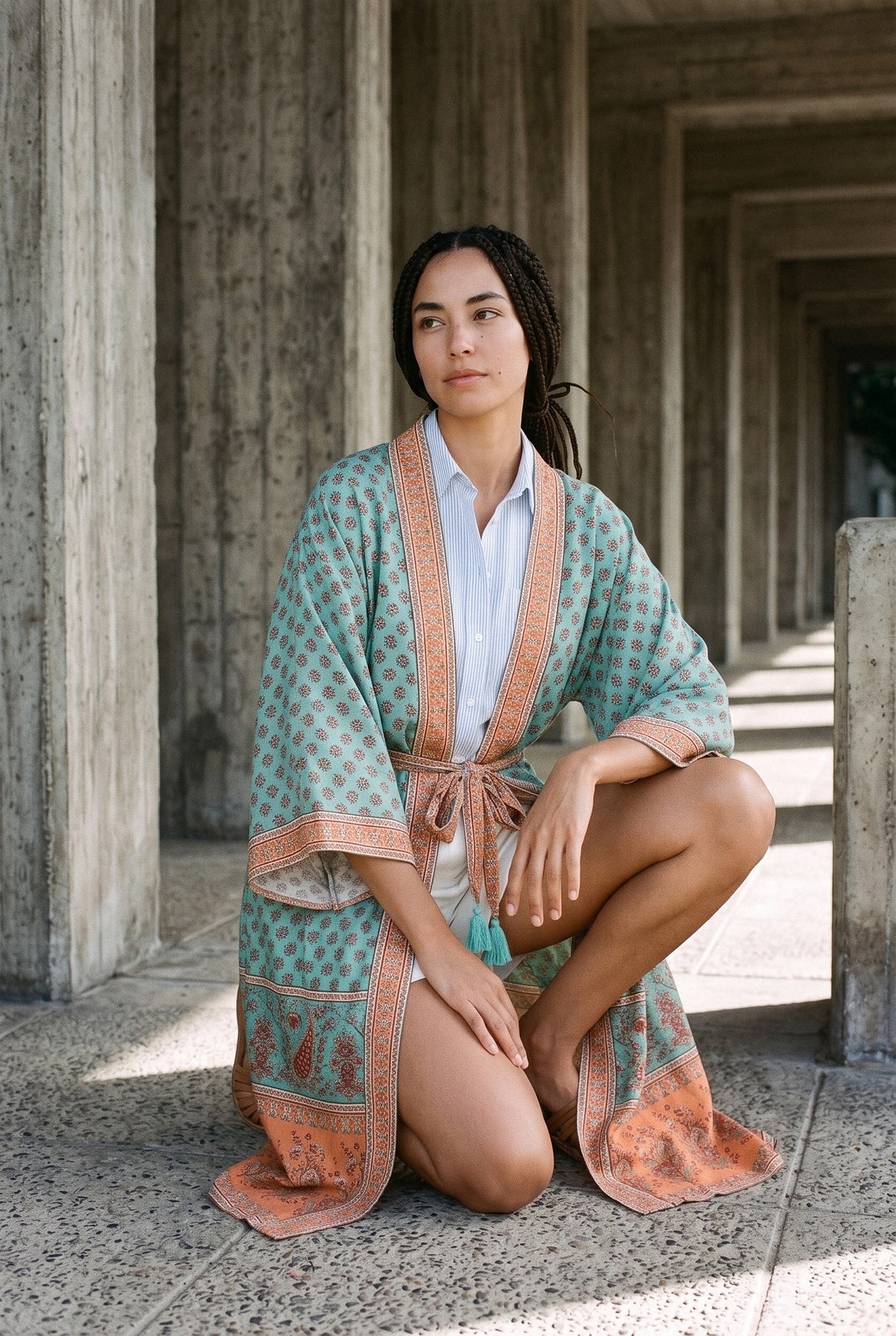 Kimono largo Nagisa, cenefas y flores, turquesa: modelo con una rodilla en el suelo, bajo una columnata brutalista de hormigón