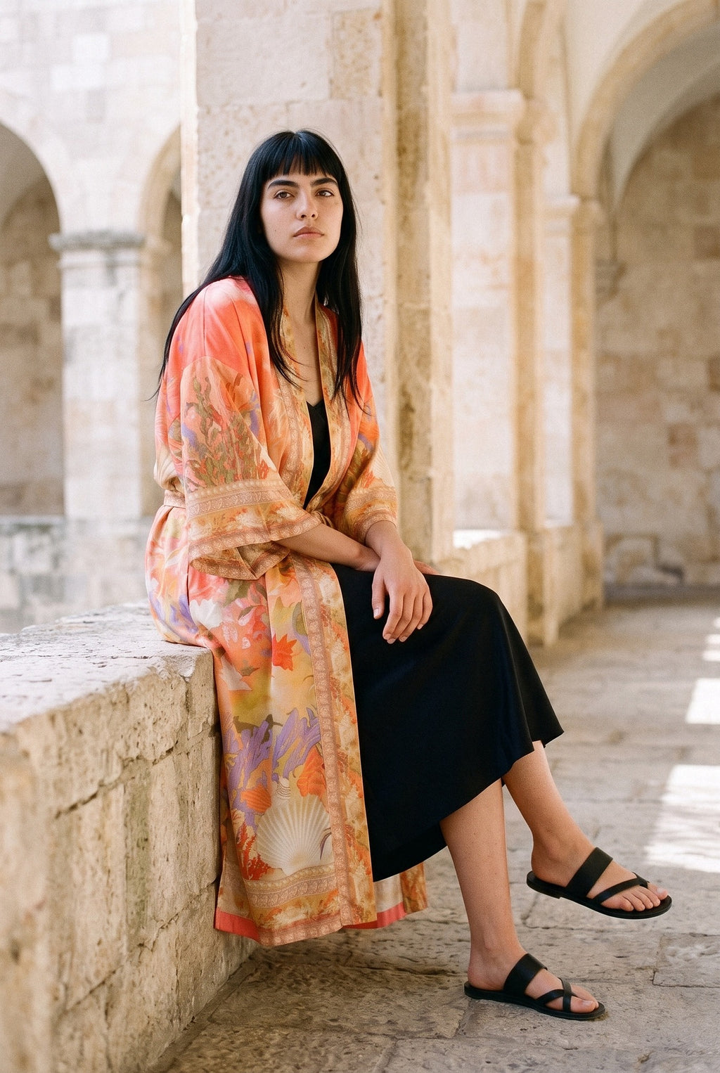 Kimono largo Sangoshō, flores, coral: modelo sentada de lado con las piernas cruzadas, en un claustro de arcos de arenisca