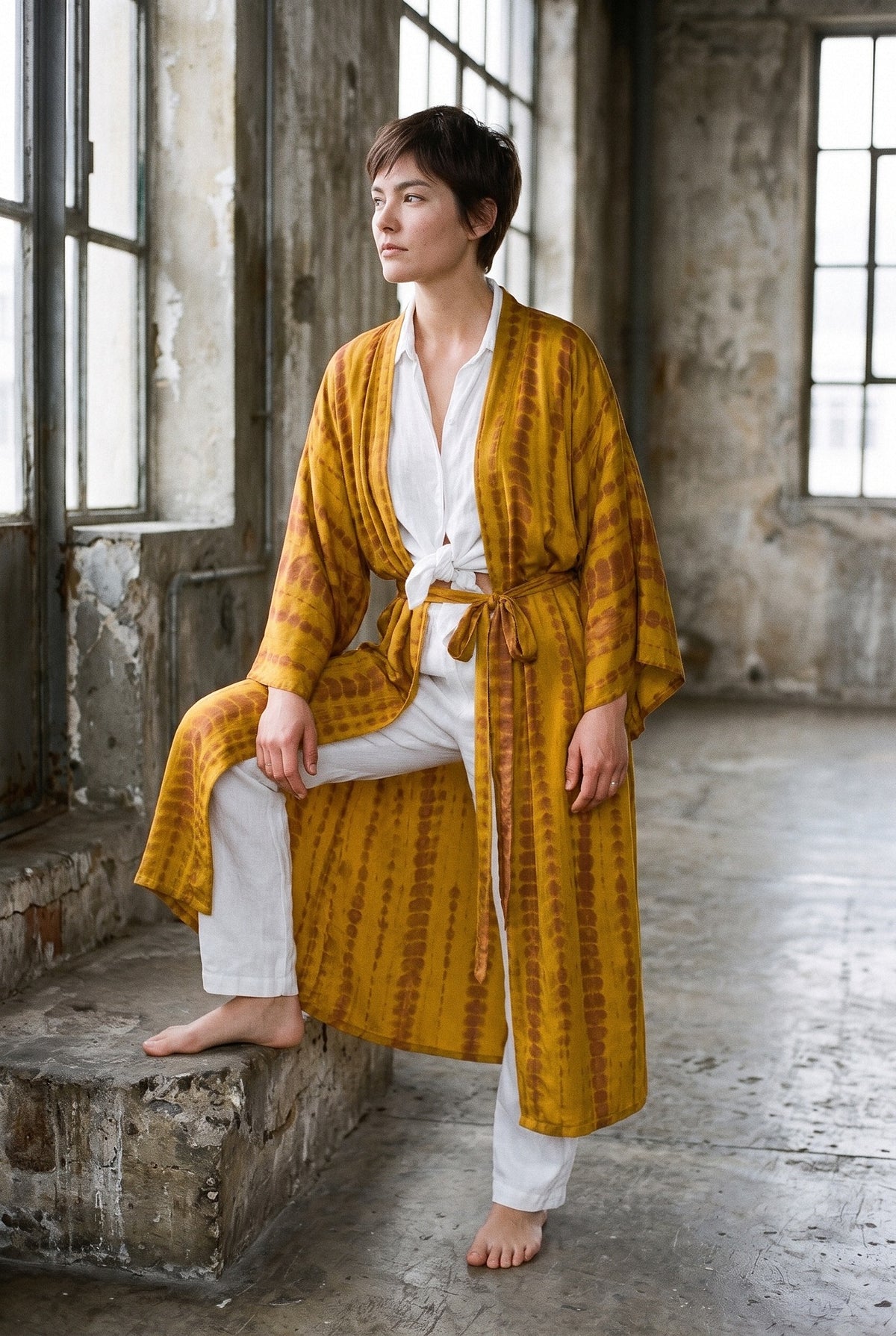 Kimono largo Shibori, efecto tie-dye, mostaza: modelo con un pie en un escalón, en un loft industrial con ventanales de acero
