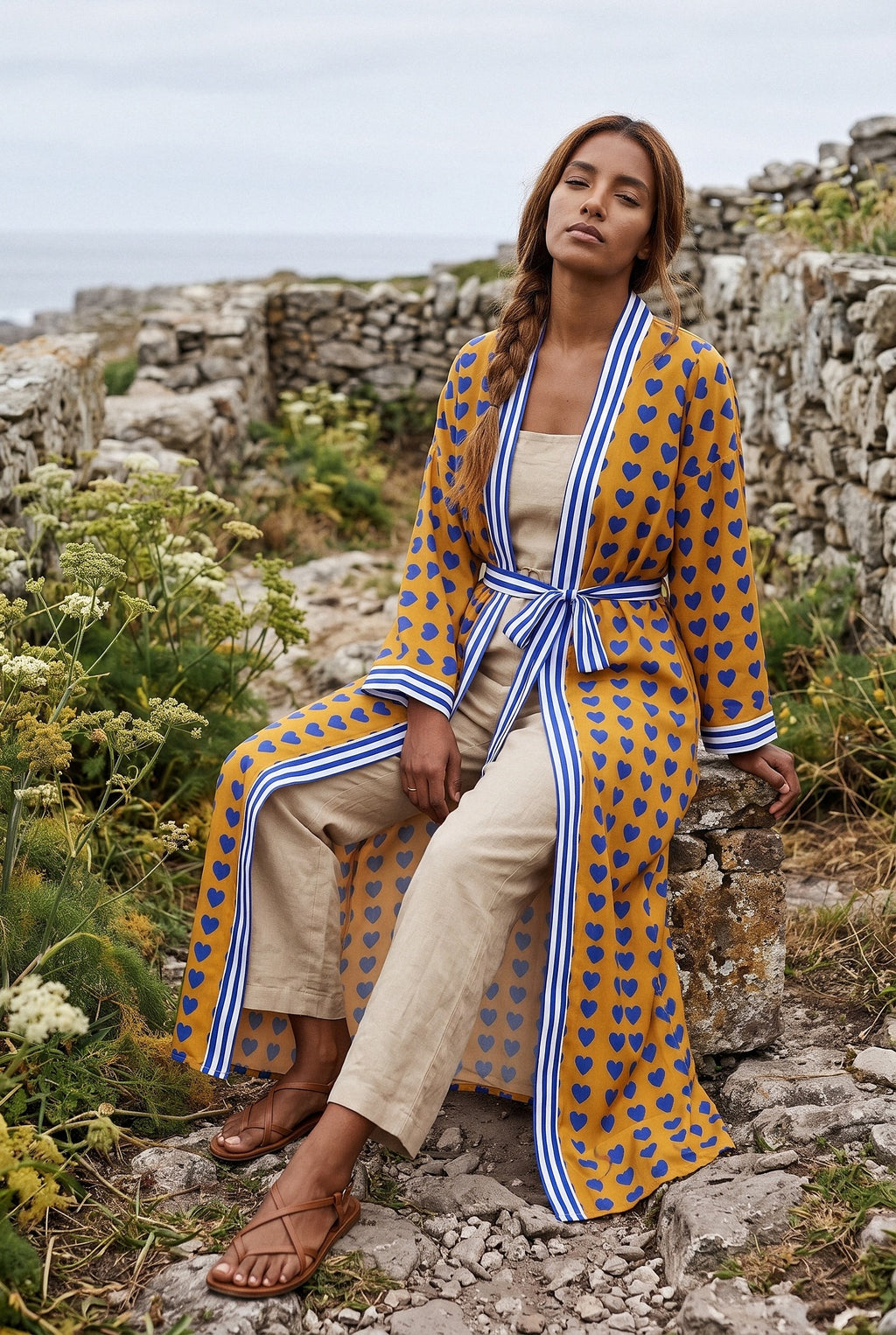 Kimono largo Shimauma, corazones azules, amarillo: modelo sentada con una pierna estirada, en un sendero costero de Galicia