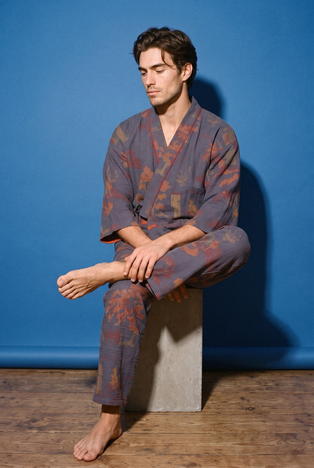 Pijama kimono Momiji, chaqueta y pantalón, azul marino: modelo sentado de lado con las piernas cruzadas, en estudio, fondo azul cobalto