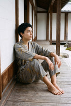Pijama kimono Momiji, chaqueta y pantalón, gris: modelo sentada contra la pared, en un jardín de grava blanca rastrillada