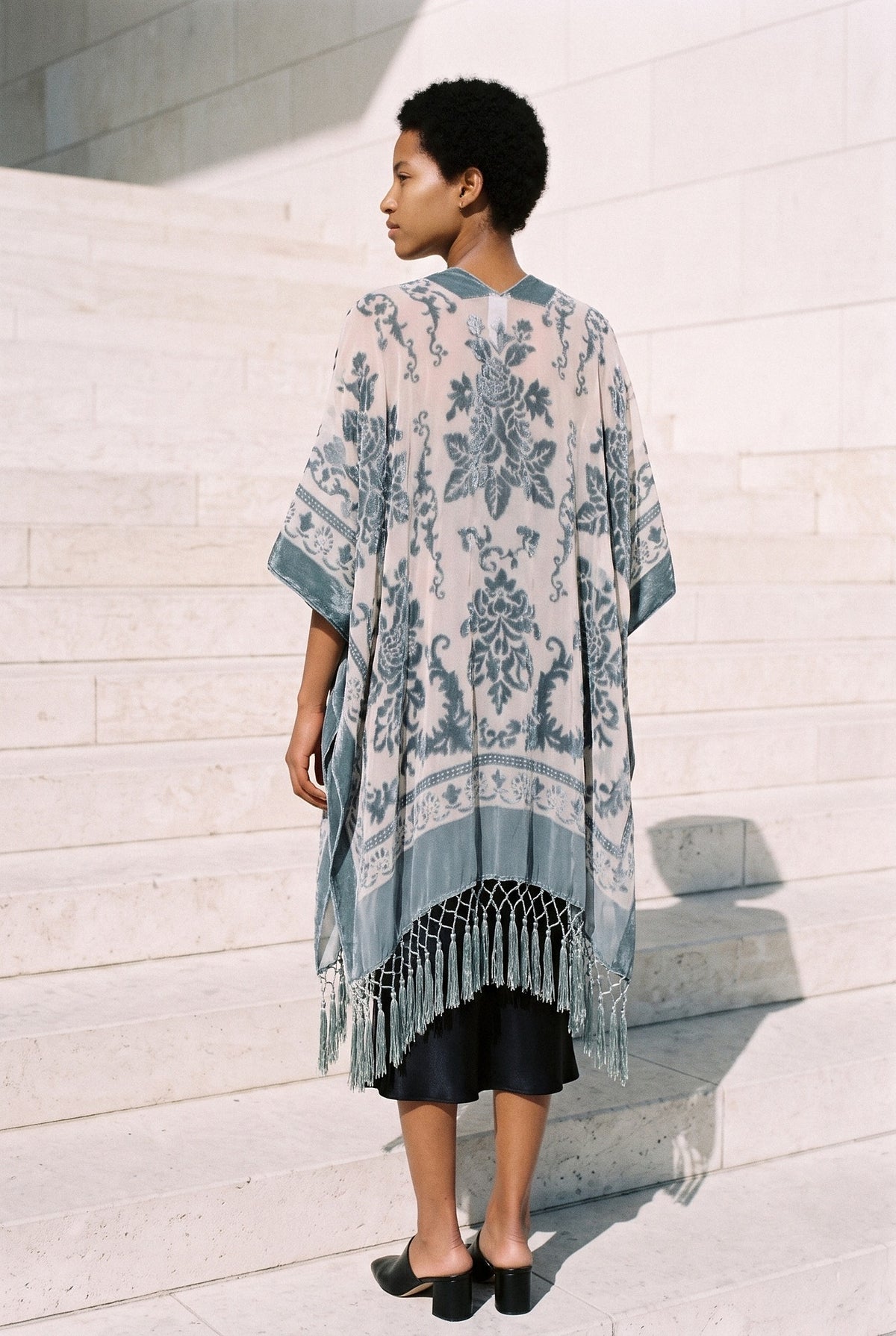 Poncho kimono Kage, terciopelo devoré, gris azulado: vista de espaldas, en las escaleras de piedra clara de un museo