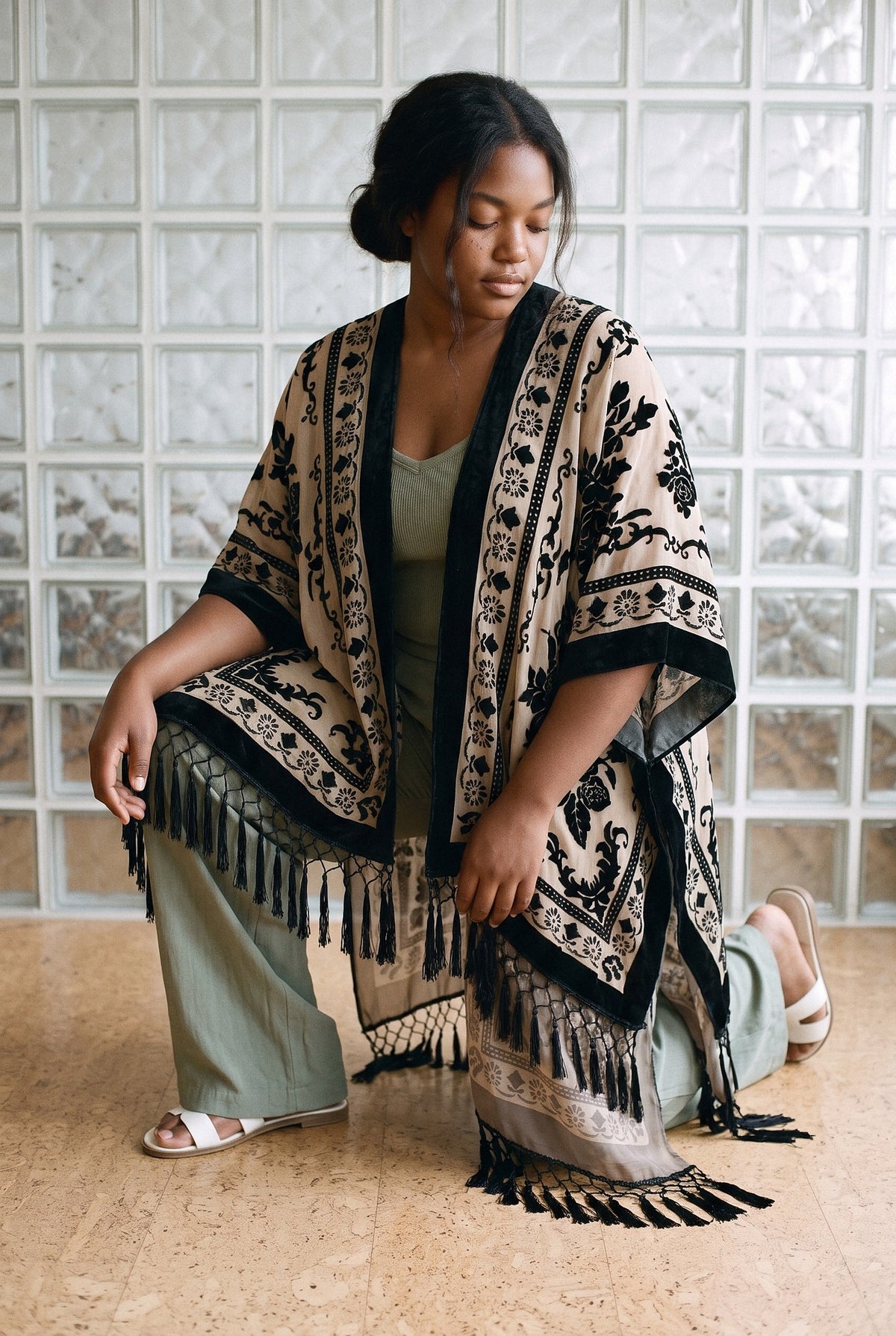 Poncho kimono Kage, terciopelo devoré, negro y crudo: modelo con una rodilla en el suelo, en un piso de los años 70 con pared de pavés
