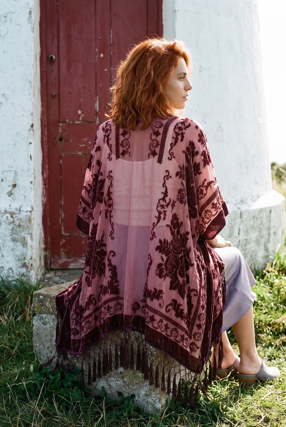 Poncho kimono Kage, terciopelo devoré, rosa antiguo: modelo de espaldas, sentada en un bloque, al pie de un faro blanco con puerta roja