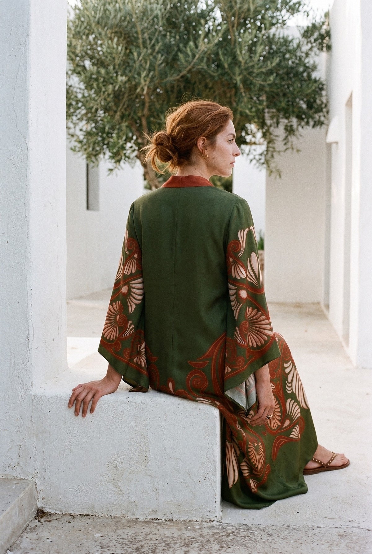 Vestido kimono Akagane, estampado verde oliva y cobre: modelo de espaldas, sentada en un bloque, junto a una casa blanca con un olivo en Ibiza